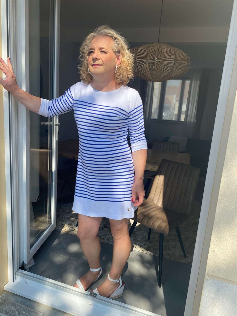 Sabine debout sur le seuil d’une porte-fenêtre, en robe blanche rayée bleu, regard tournée vers la lumière, image illustrant un parcours de transidentité tardive, de questionnement de genre et d’acceptation de soi.